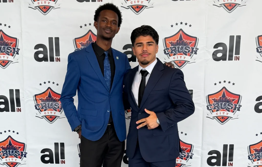 Two men in formal attire pose confidently in front of a branded backdrop featuring "Crush City" and "All In.