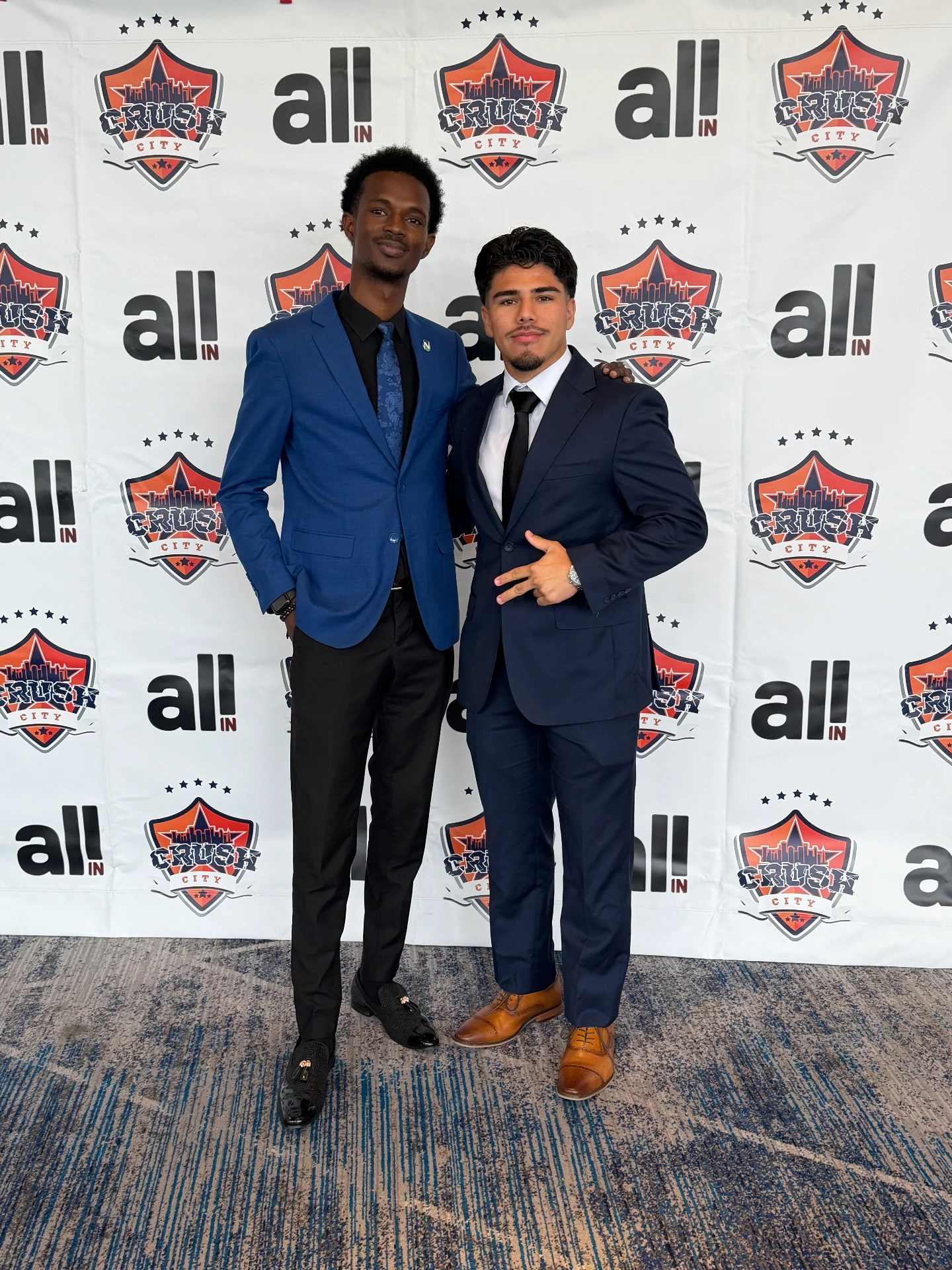 Two men in formal attire pose confidently against a branded backdrop at a promotional event.